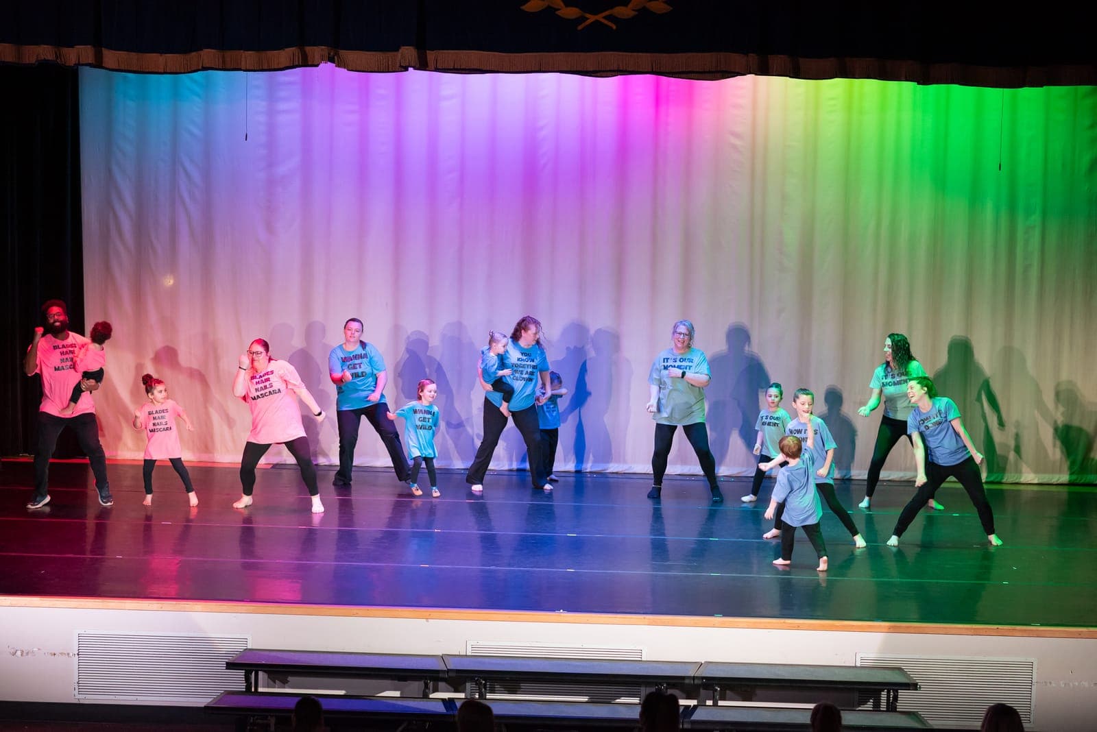 A large all-ages hip-hop group in pink and blue graphic tees strikes an energetic pose across the stage in front of a rainbow-lit curtain.