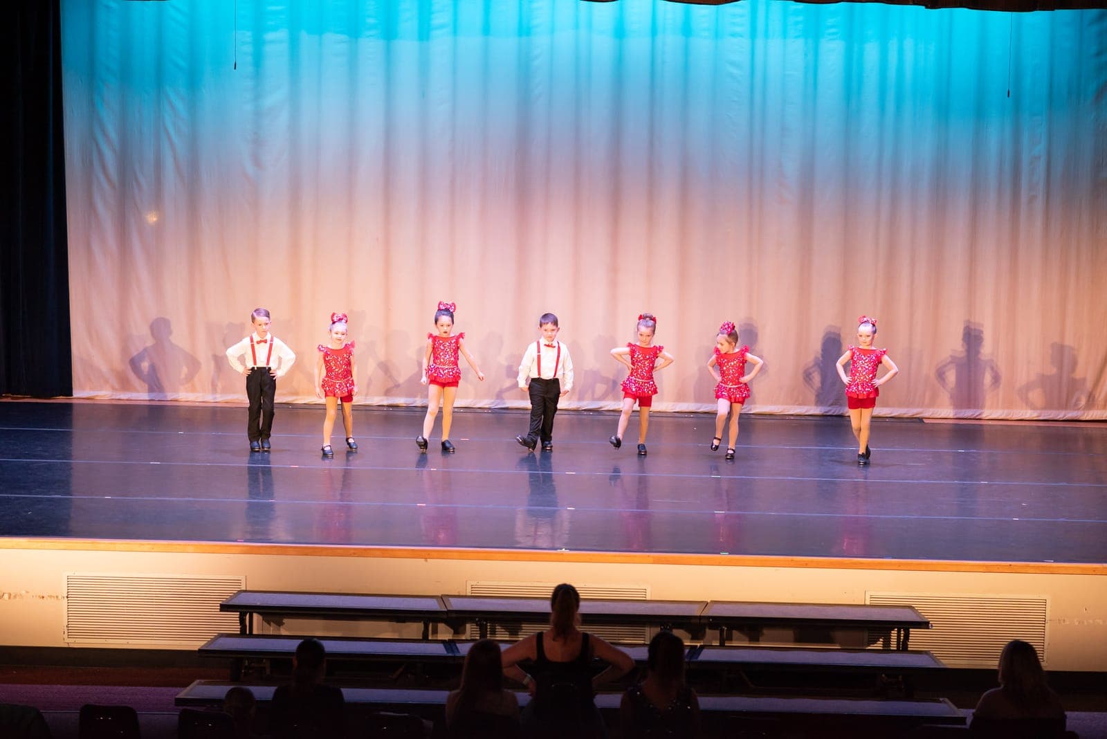 Young tap dancers line up across the stage — girls in red sequined dresses and boys in white shirts with red suspenders — before a teal curtain.