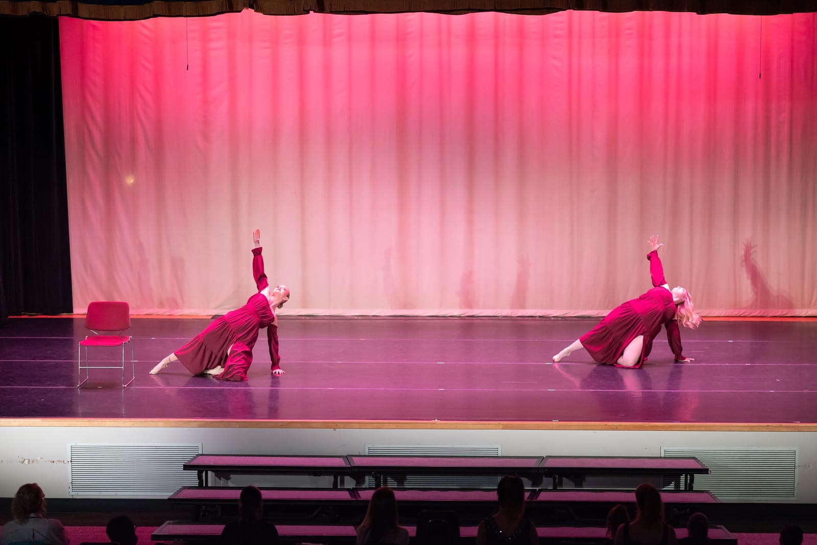 Two lyrical dancers in long burgundy dresses arch into expressive backbends on a pink-lit stage, one beside an empty chair.