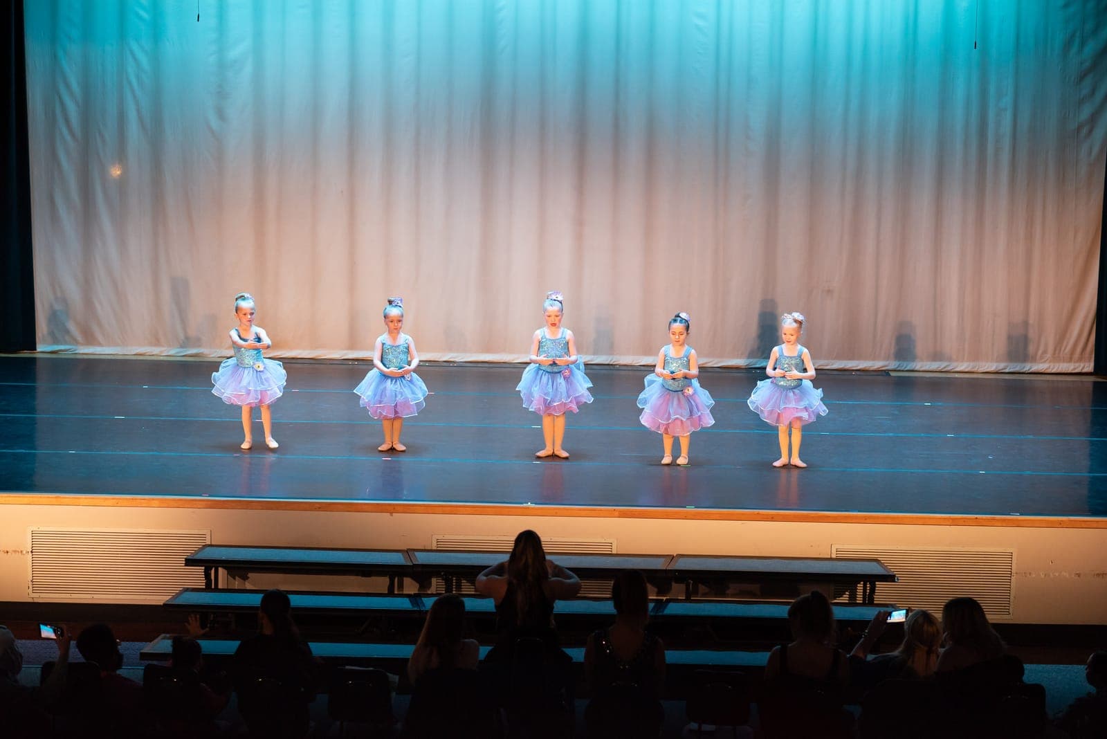 Five young ballerinas in sparkly blue tutus stand poised in a row before a softly lit teal curtain.