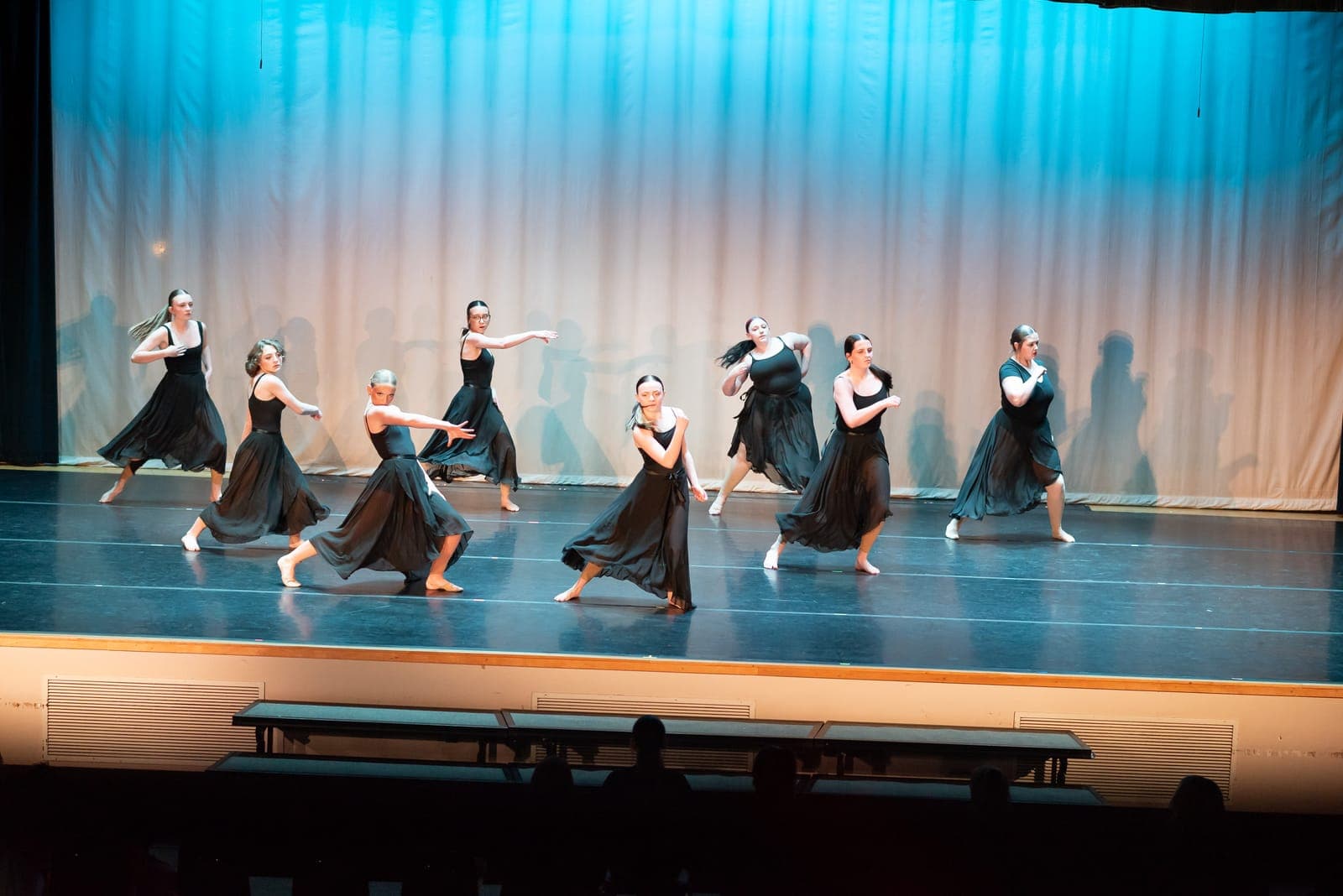 A contemporary ensemble in flowing black dresses lunges through a synchronized phrase under cool blue stage light.