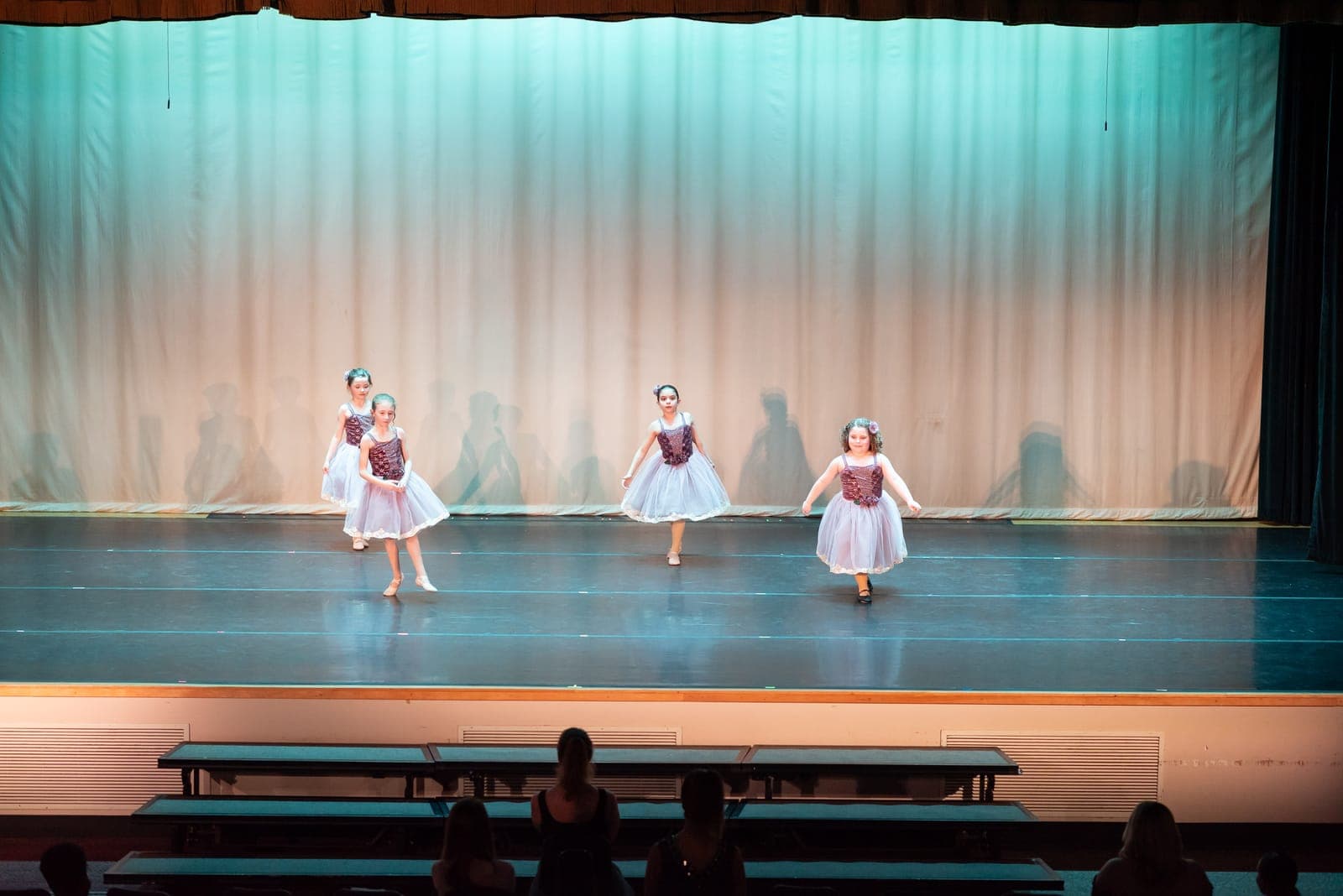 Four young ballet students in romantic lilac tutus dance forward together in front of a green-lit curtain.