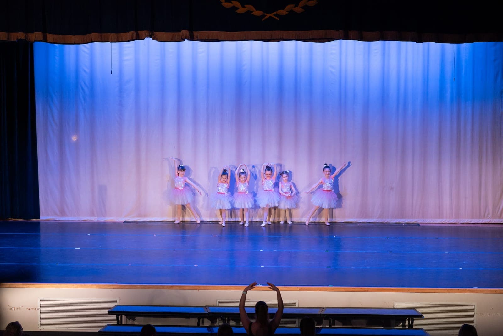 Six little ballerinas in pale blue tutus pose with arms framed overhead under soft blue stage lighting.