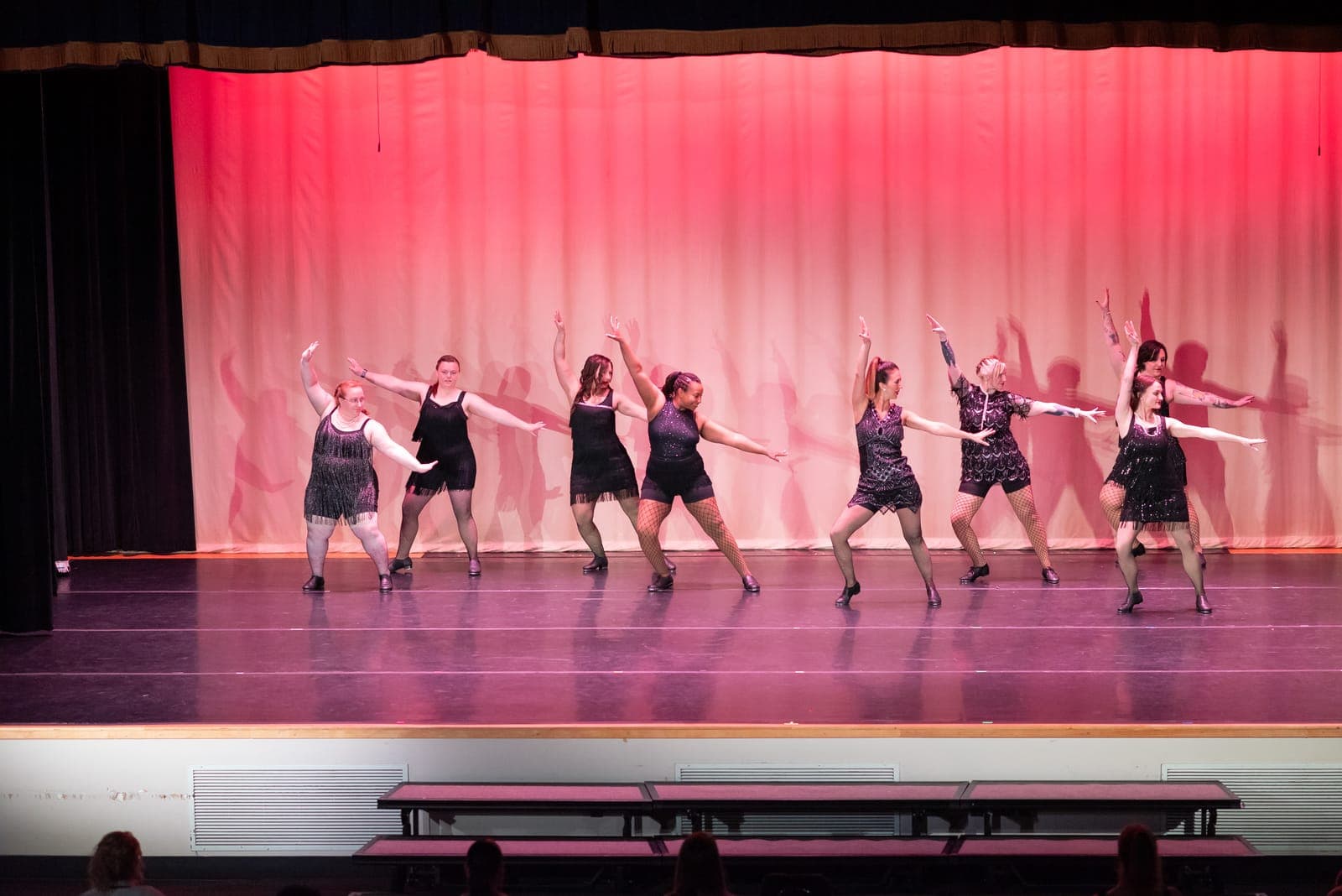 Adult tap dancers in black fringed flapper dresses and fishnets hit a sharp jazz-hands pose against a red-lit curtain.