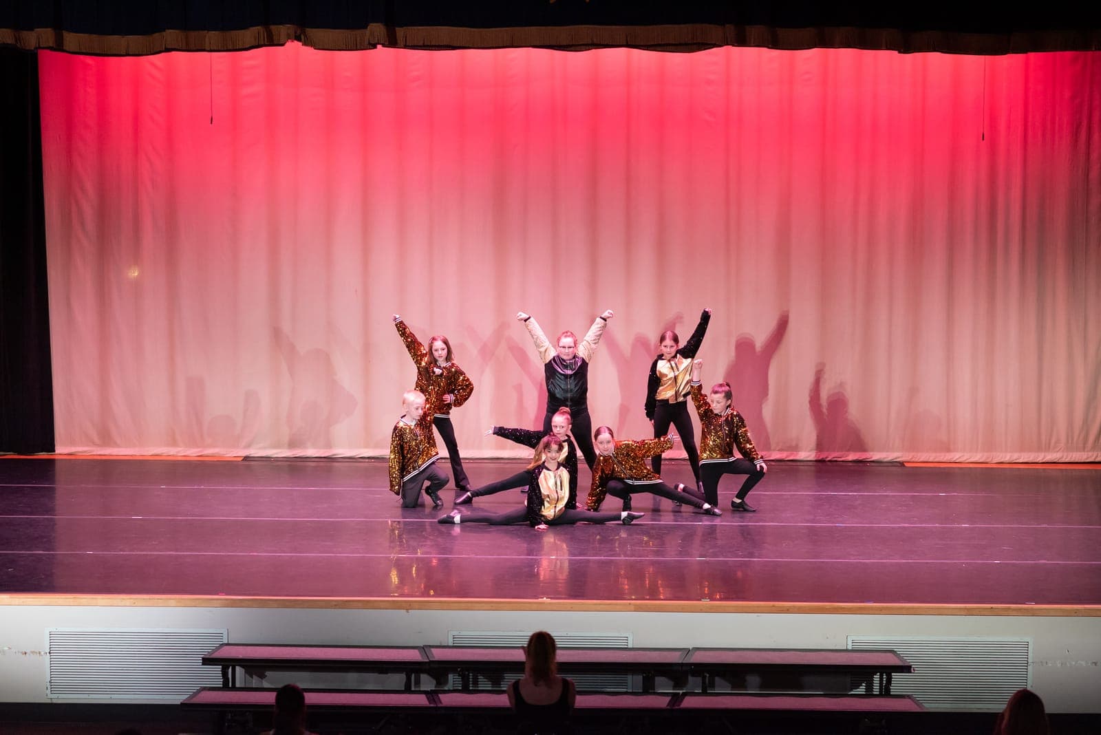 A hip-hop troupe in gold sequined jackets holds a dynamic group pose, one dancer dropping into a center split, before a red-lit curtain.