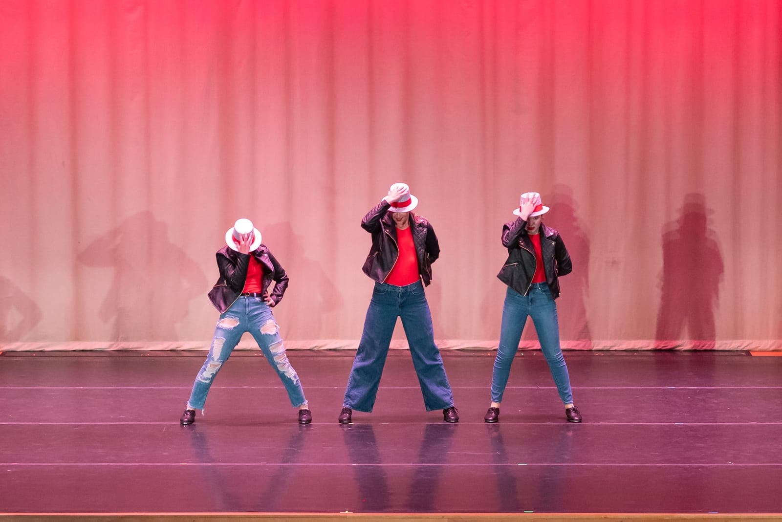 Three jazz dancers in leather jackets, red tops, and white fedoras tip their hats in unison on a red-lit stage.