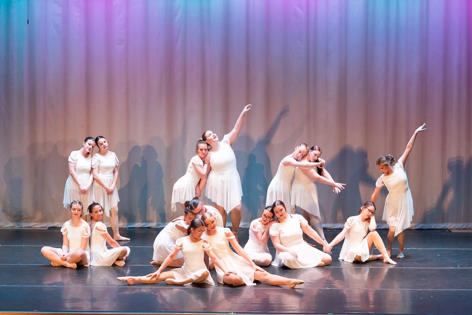 A large lyrical ensemble in flowing white dresses melts into an interlocked group tableau under purple and blue light.