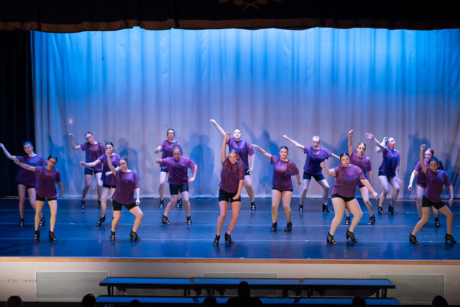 A big jazz company in purple mesh tops and black shorts powers through a synchronized routine before a blue-lit curtain.